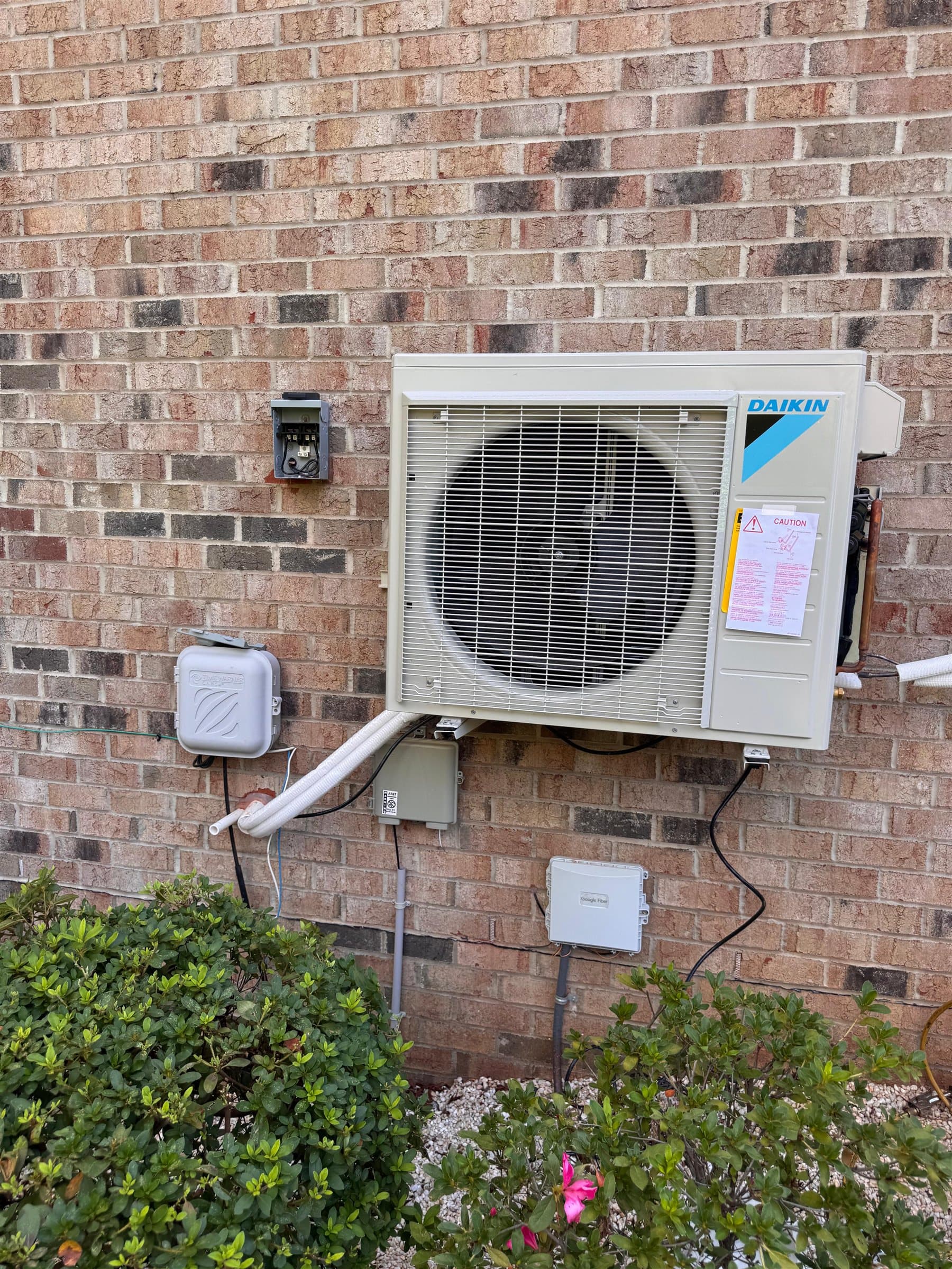 Daikin ductless mini-split condenser mounted on a brick wall above the shrubs, with its line set running through a white cover.