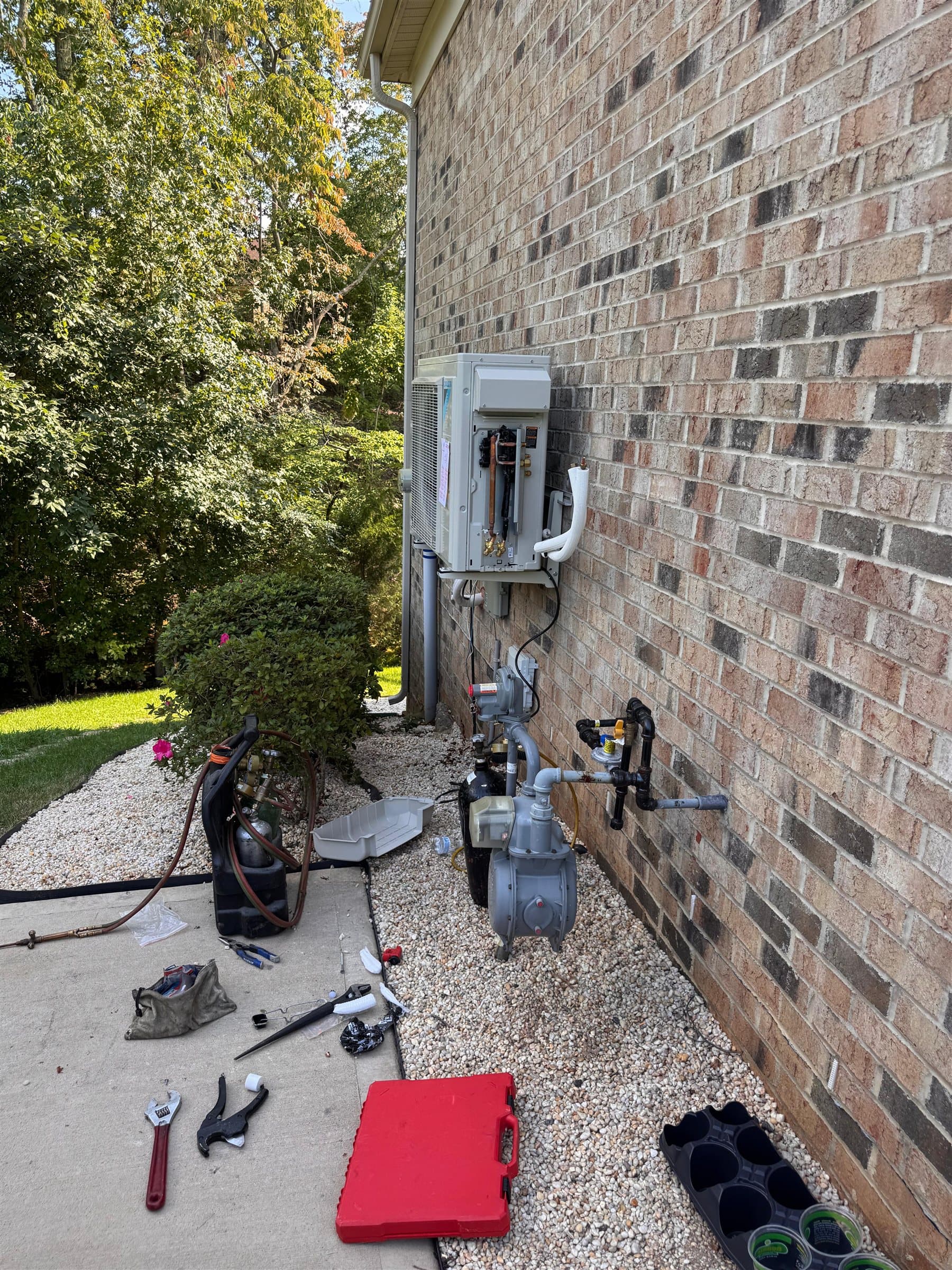Ductless mini-split outdoor unit on a brick wall, with a vacuum pump hooked to the line set and tools laid out on the walkway.