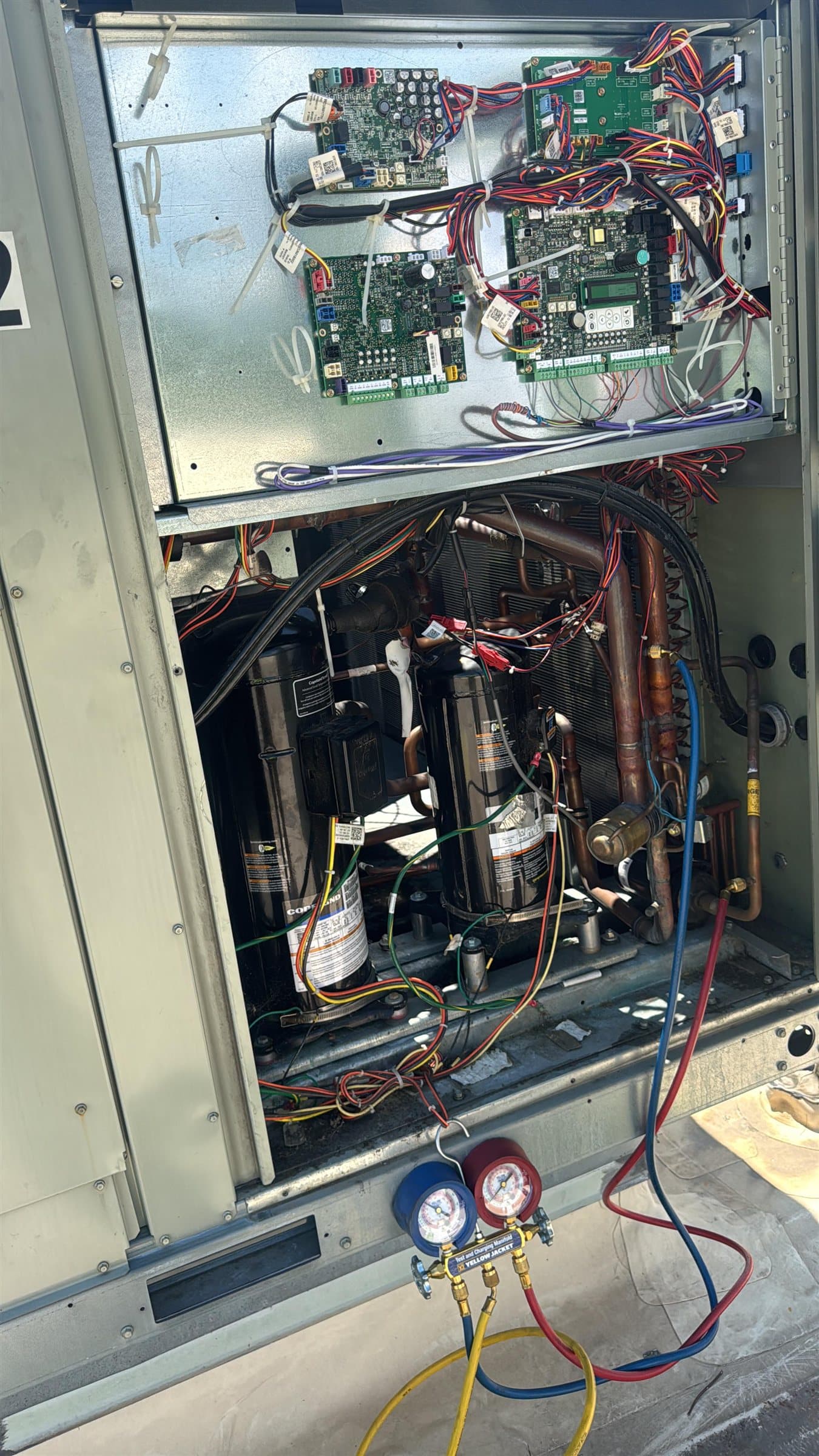 Commercial rooftop unit with its service panel open, showing two compressors and control boards, with a manifold gauge set connected to the refrigerant lines.